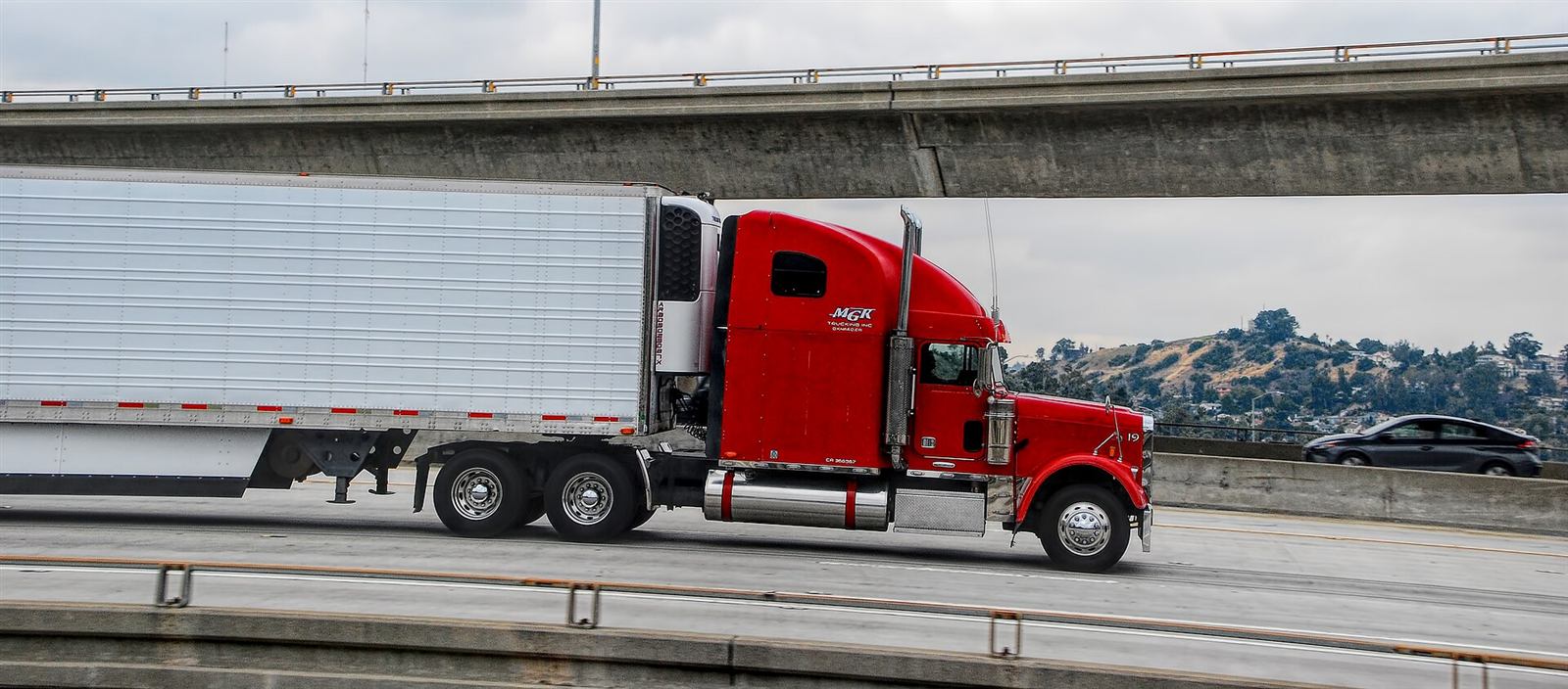 A red semi-truck hauling a trailer under a highway overpass